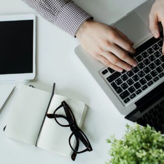 Hands working on a laptop with a notebook and tablet beside it.