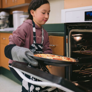 Child wearing apron and oven mitts places a pizza in the oven.