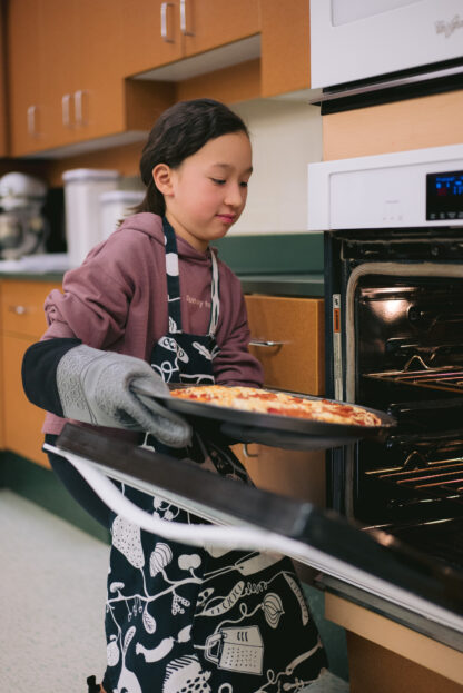 Child wearing apron and oven mitts places a pizza in the oven.