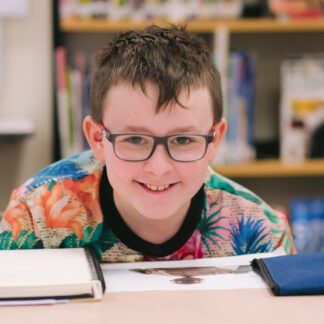 A young boy in glasses smiles at the camera while sitting over the top an open sketchbook.