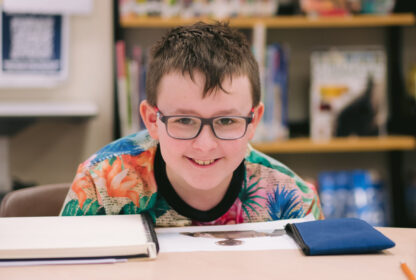 A young boy in glasses smiles at the camera while sitting over the top an open sketchbook.