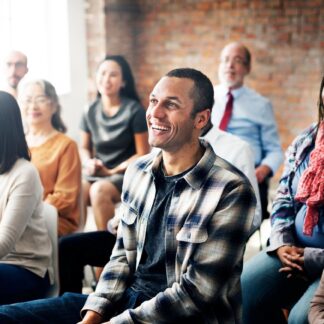 A group of adults sit in multiple rows of chairs laughing at something in front of them.