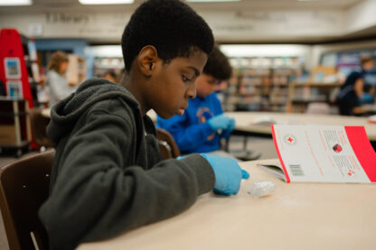 A young person wearing medical gloves practices unrolling gauze in a classroom with a booklet bearing the Red Cross logo in front of him.