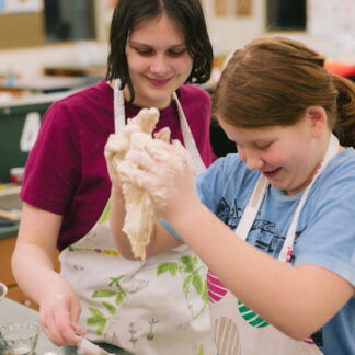 Two children smile as they work with pizza dough in a kitchen.