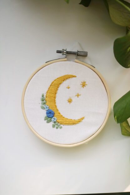 An embroidery hoop holds a design of a crescent moon with flowers and stars added to the design.