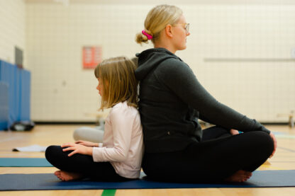 A mom and daughter sit back to back and crosslegged on a yoga mat with eyes closed in a meditative pose.