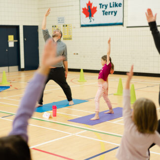 A barefoot group of parents and their children extend there arms upwards as they each stand on their yoga mats.