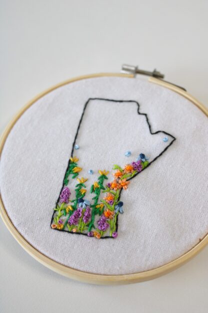 An embroidery hoop with an outline of the province of Manitoba with embroidered flowers added to the bottom.
