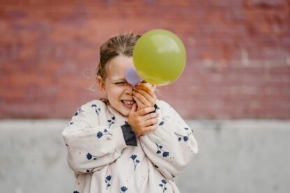 Child holding a balloon up to their face standing in front of a brick wall.