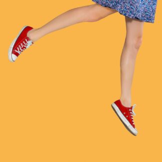 The legs of a young woman wearing a mini skirt and red sneakers are seen against a yellow background