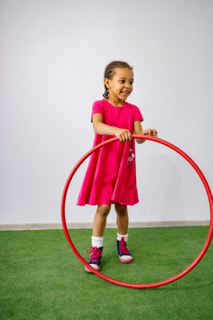 A young girl in a red dress smiles as she plays with a hula hoop.