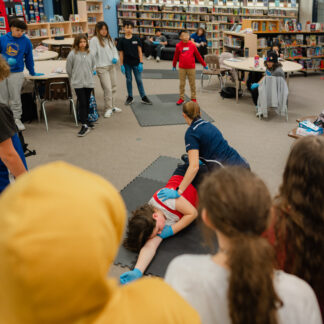 A group of students observe as an instructor demonstrates on another student laying on the floor of a CPR class