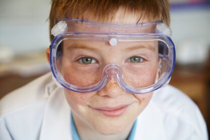 A child wearing safety glasses and a lab coat grins at the camera